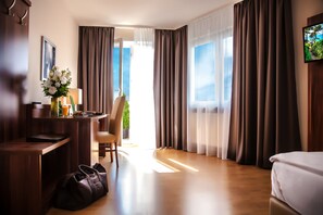A hotel room with a bed, a desk with a chair, a small table with a vase of flowers, and a large window with curtains.