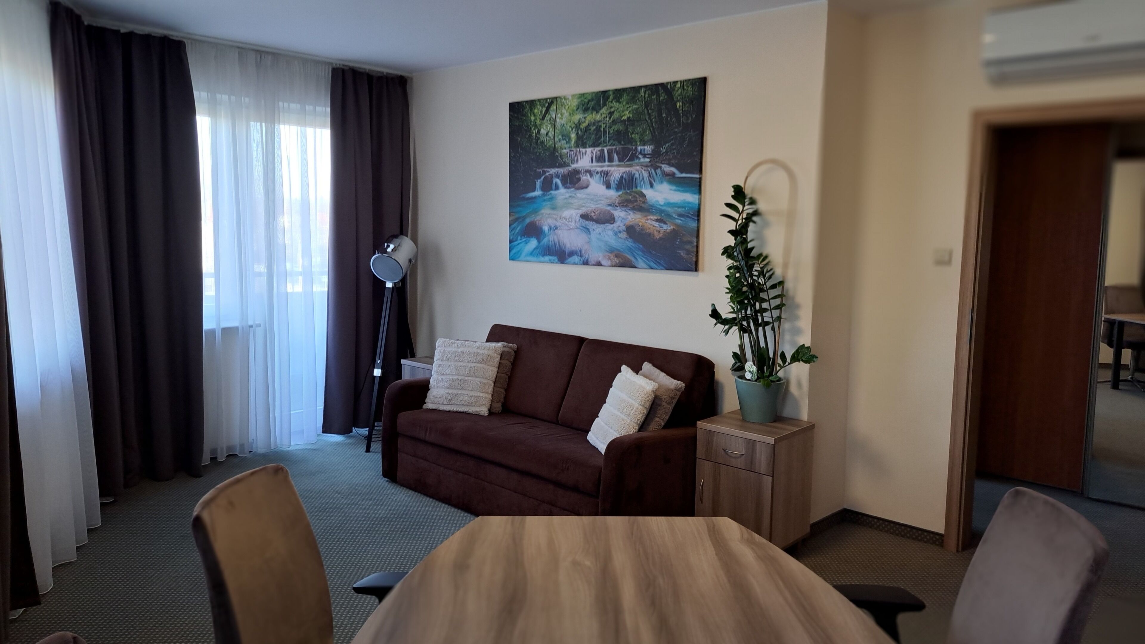 A hotel room with a brown sofa, a small wooden table, a potted plant, a floor lamp, and a painting of a waterfall on the wall.
