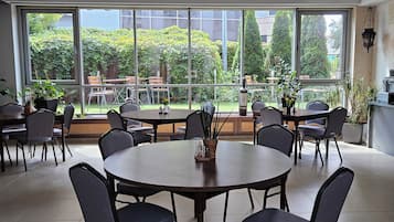 A spacious indoor area with multiple round tables and chairs, large windows offering views of a garden and buildings, and a ceiling with recessed lighting.
