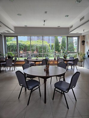 Un vaste espace intérieur doté de plusieurs tables et chaises rondes, de grandes fenêtres offrant une vue sur un jardin et des bâtiments, et d’un plafond équipé d’un éclairage encastré.