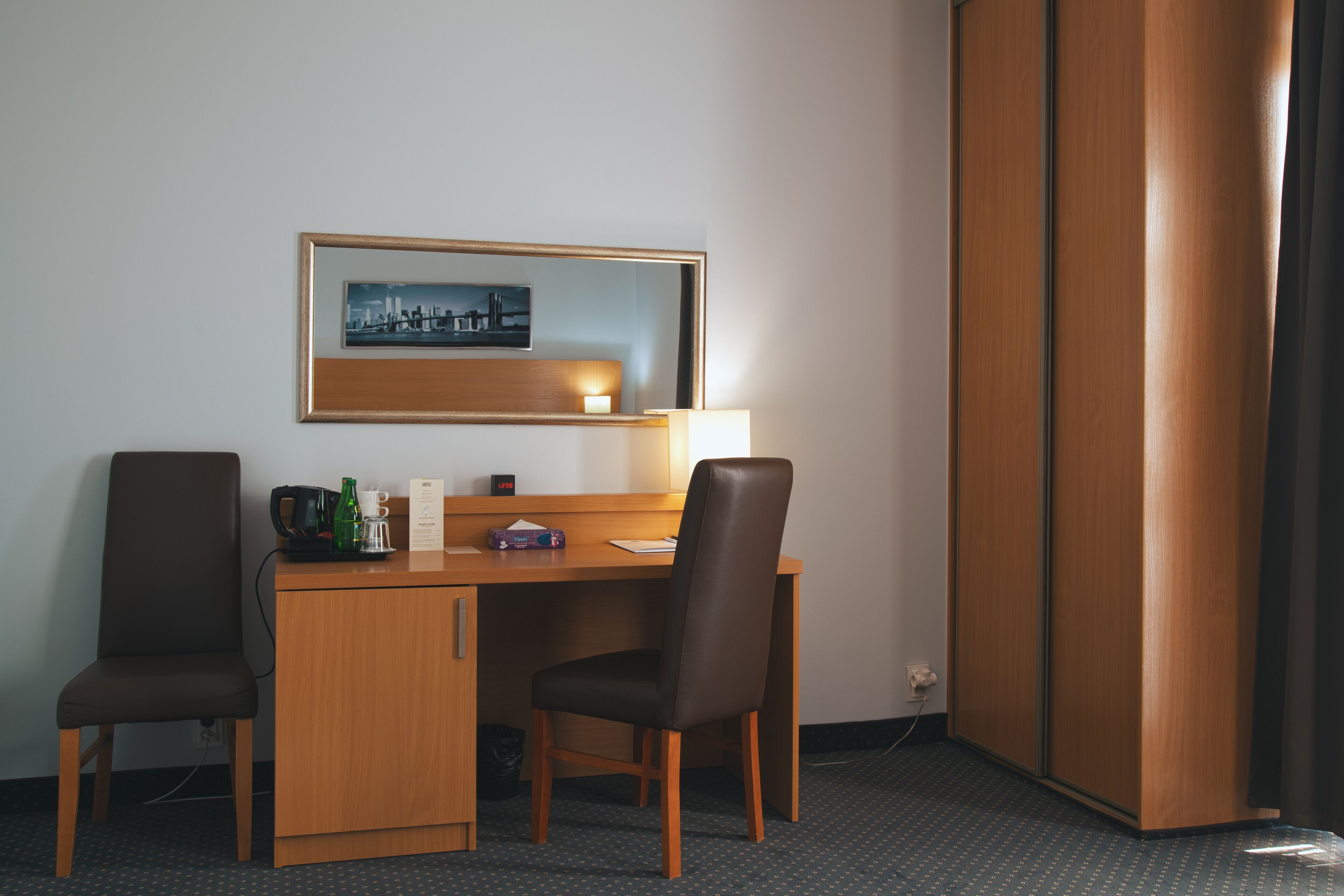A hotel room with a wooden desk, two chairs, a framed cityscape picture, a lamp, and a window with curtains.