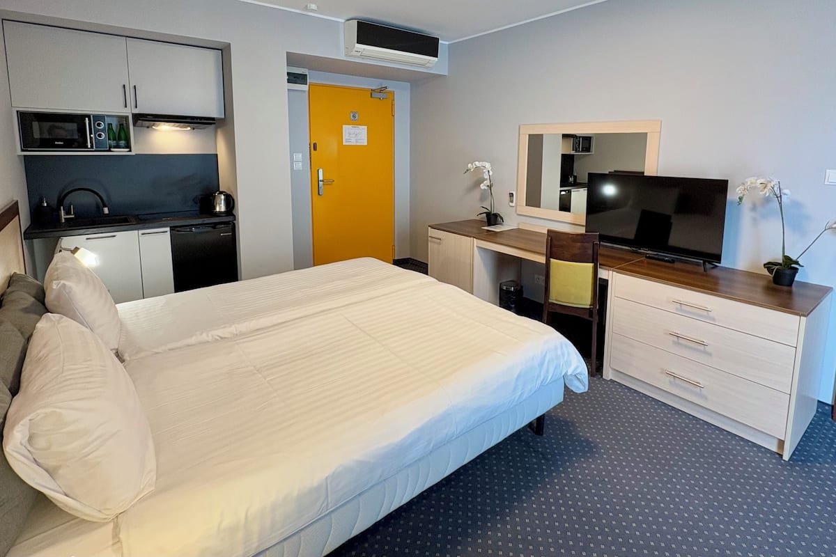 Quarto de hotel com cama, escrivaninha com televisão, cozinha compacta e banheiro.