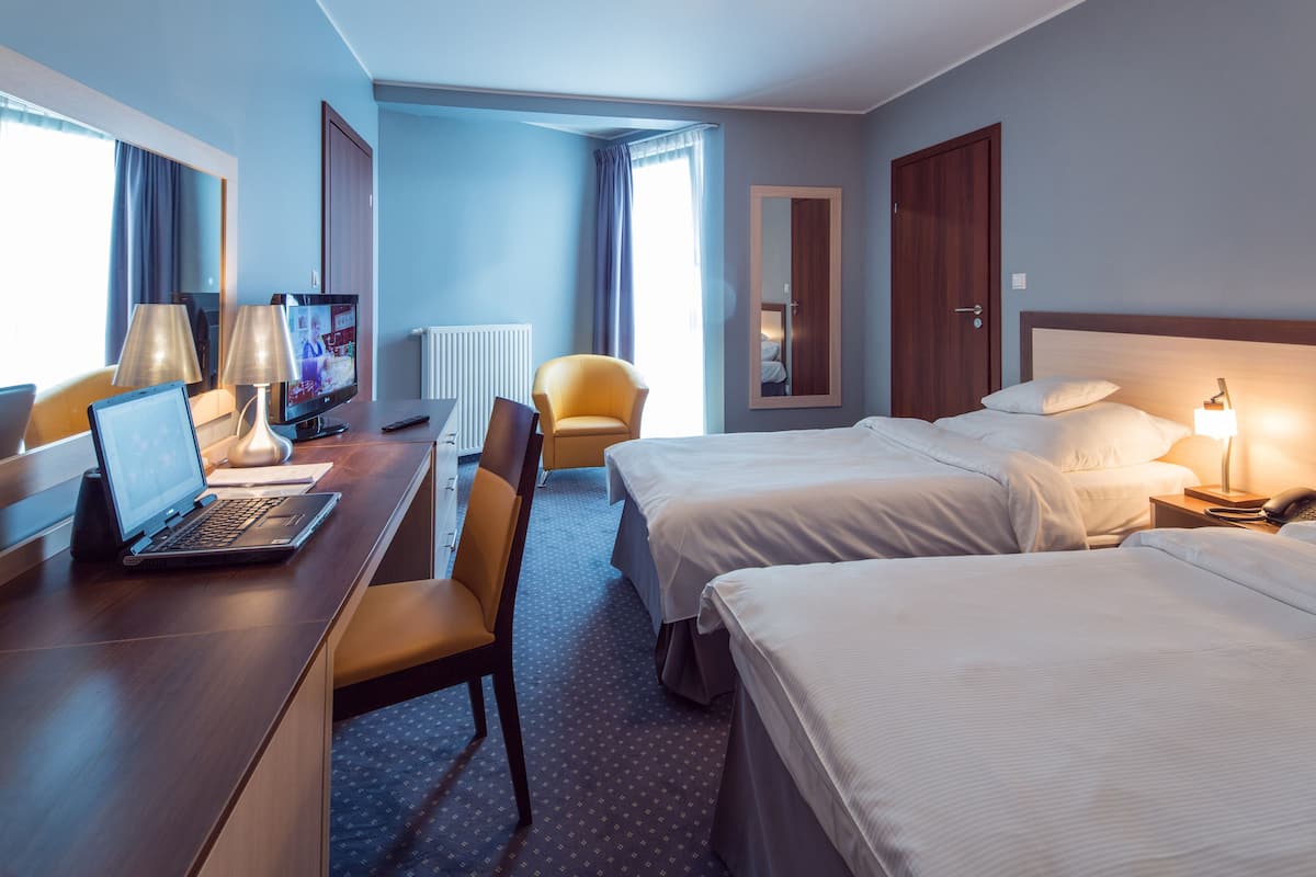 Quarto de hotel com escrivaninha, duas camas, uma cadeira, televisão e janela com cortinas.