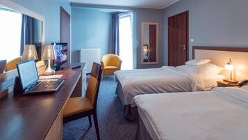 A hotel room with a desk, two beds, a chair, a TV, and a window with curtains.