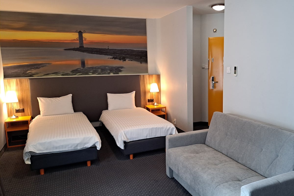 Quarto de hotel com duas camas de solteiro, um sofá cinzento, um quadro de um pôr do sol e uma porta de madeira.