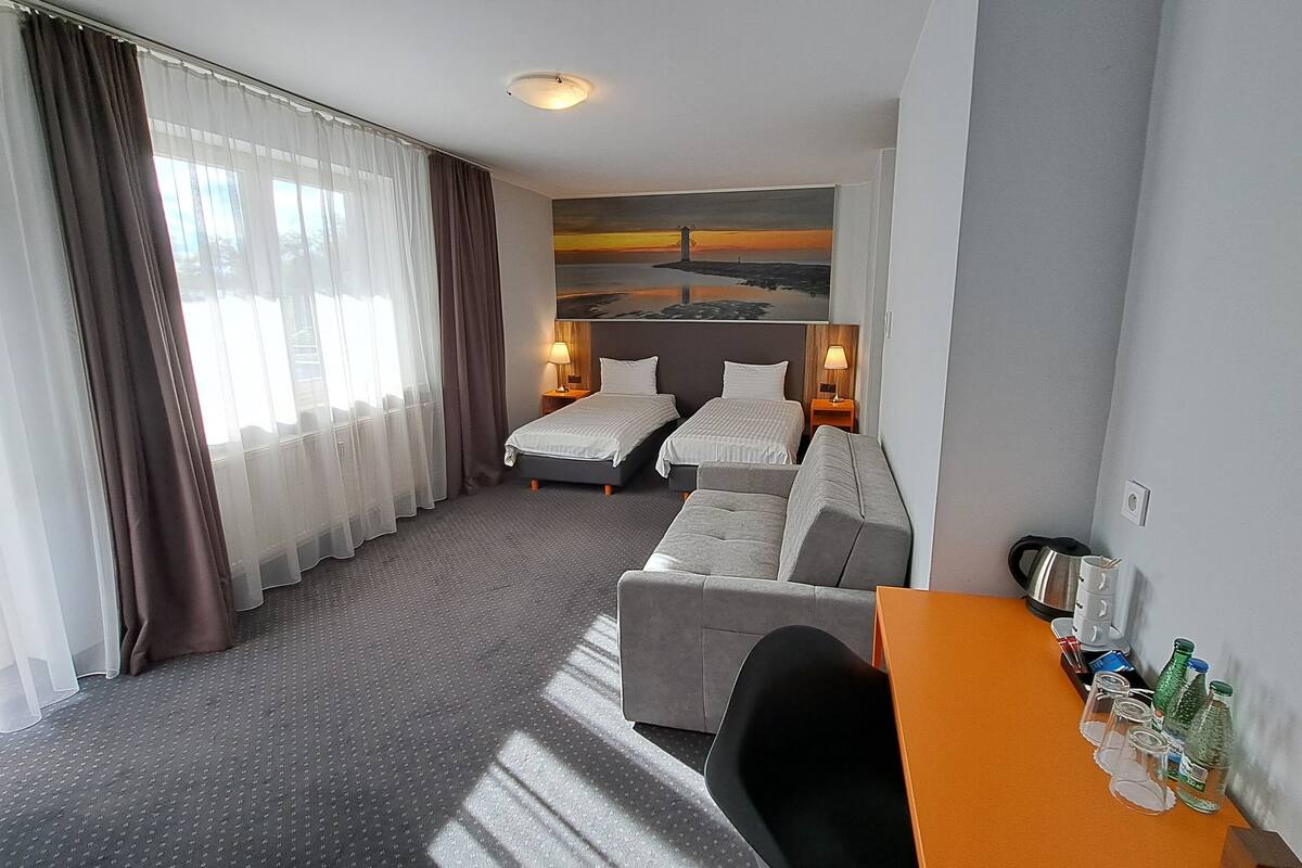 Quarto de hotel com duas camas, um sofá, uma mesa com chaleira e um quadro de um pôr do sol.