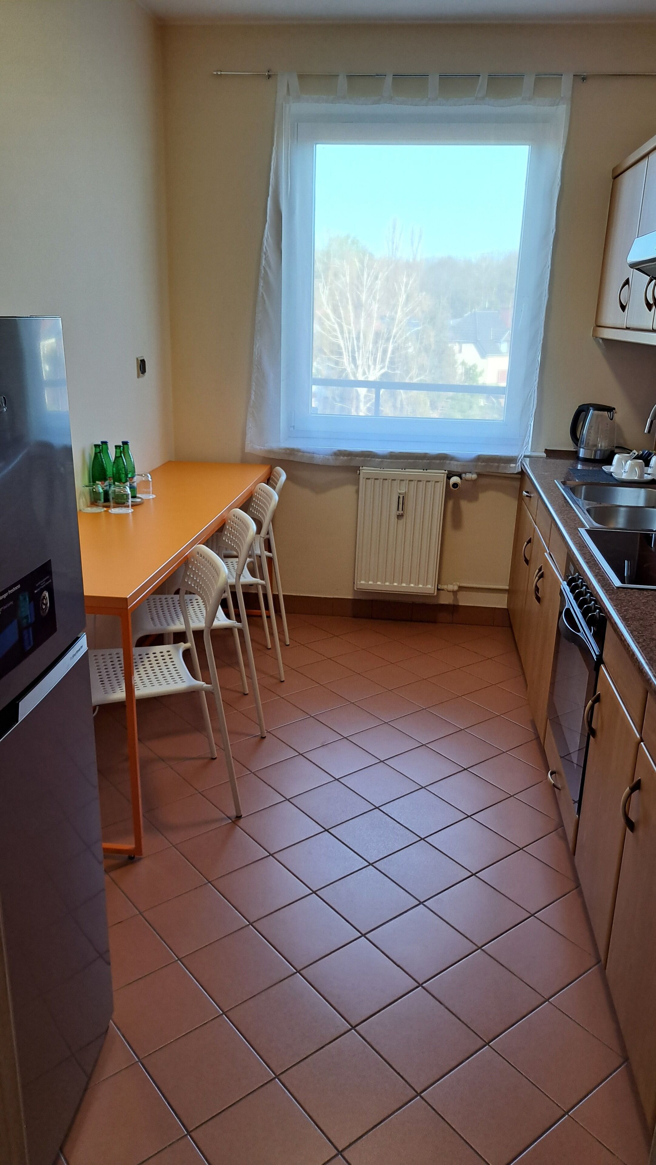 A compact kitchen with a dining table, chairs, a refrigerator, a window with curtains, and a radiator.