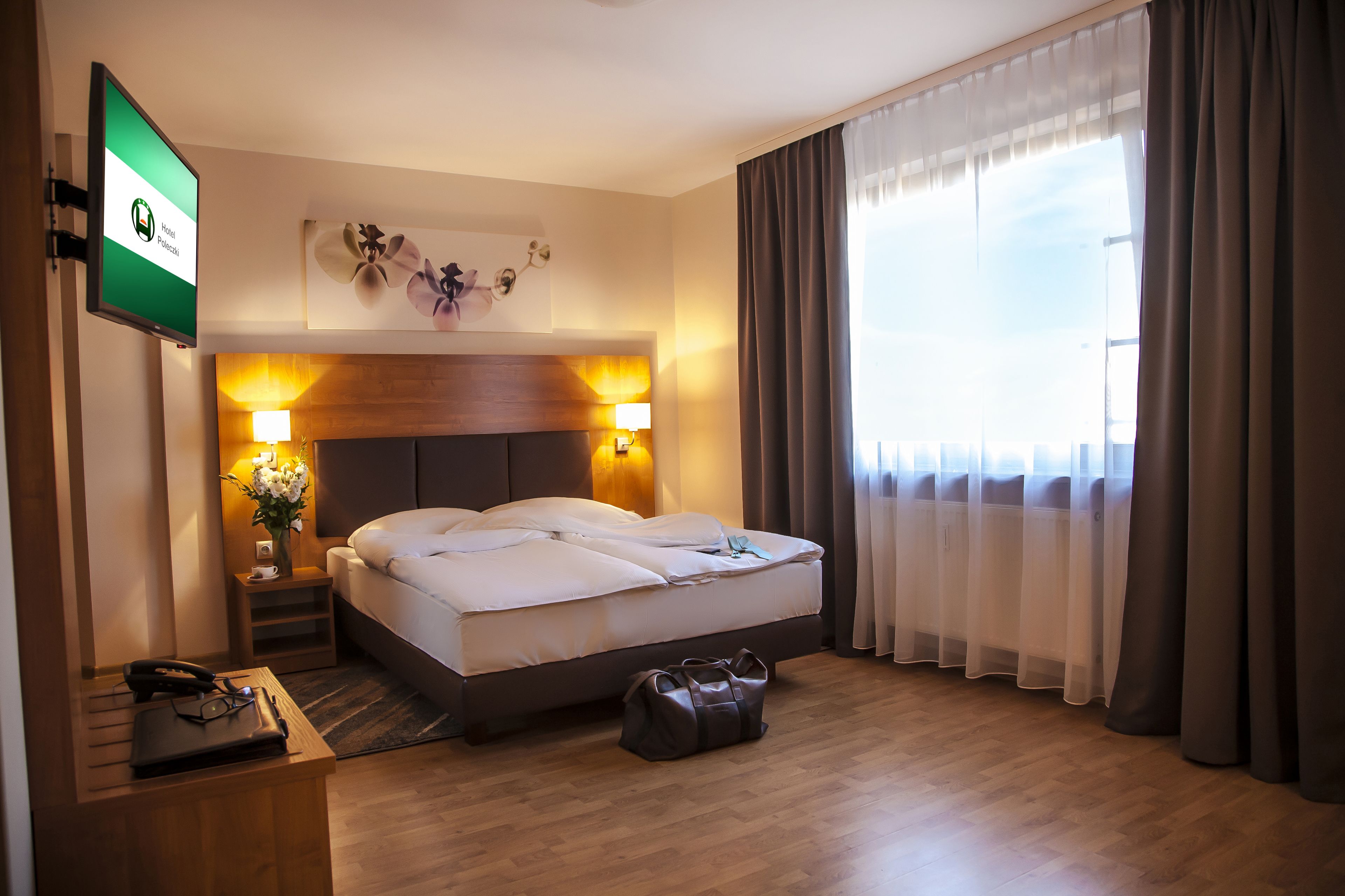 Una habitación de hotel con una cama, lámparas de noche, un televisor, un escritorio con teléfono y gafas, y un bolso en el suelo.