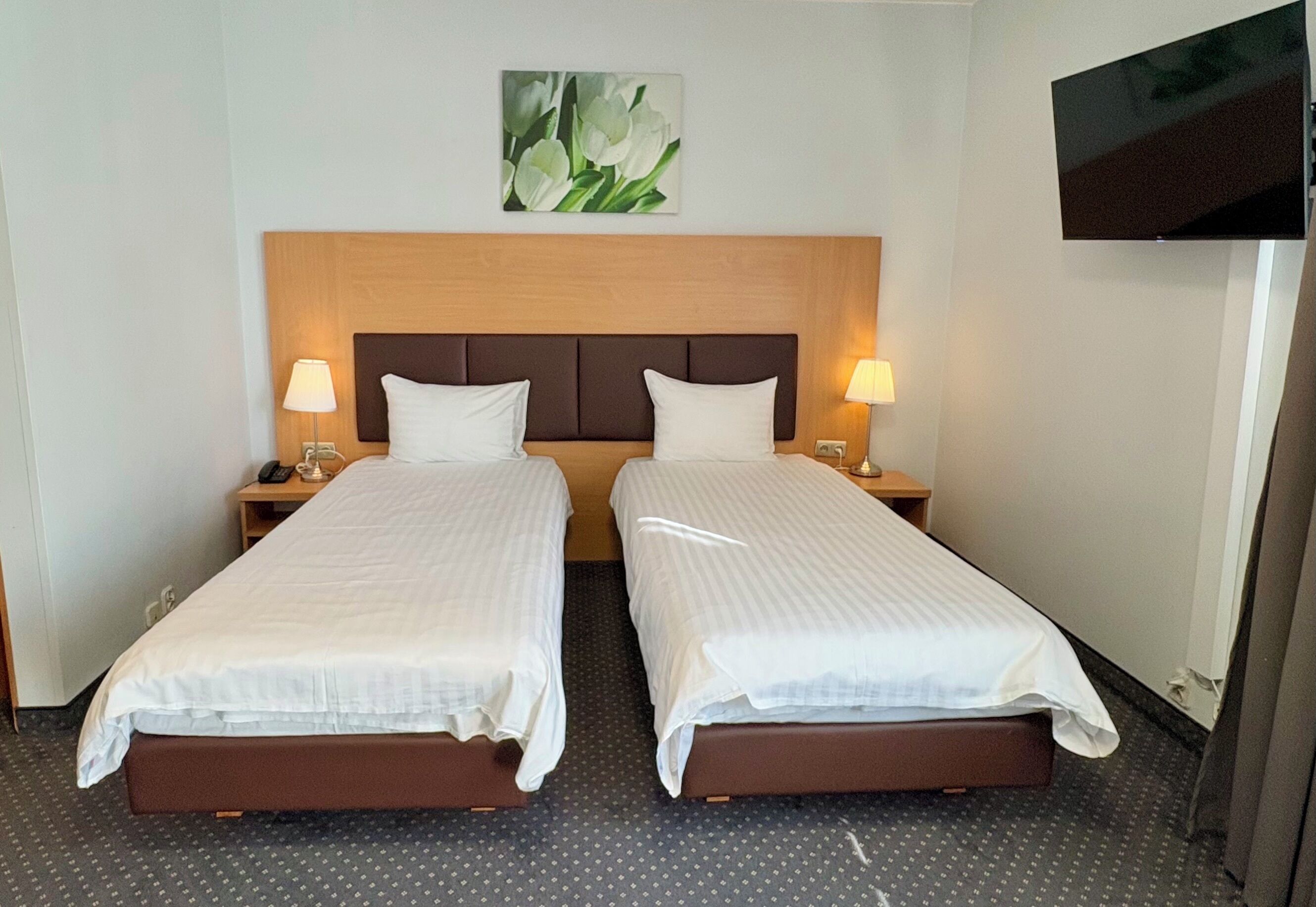 A hotel room with two single beds, a wooden headboard, a flat-screen TV, and a wall-mounted painting of flowers.