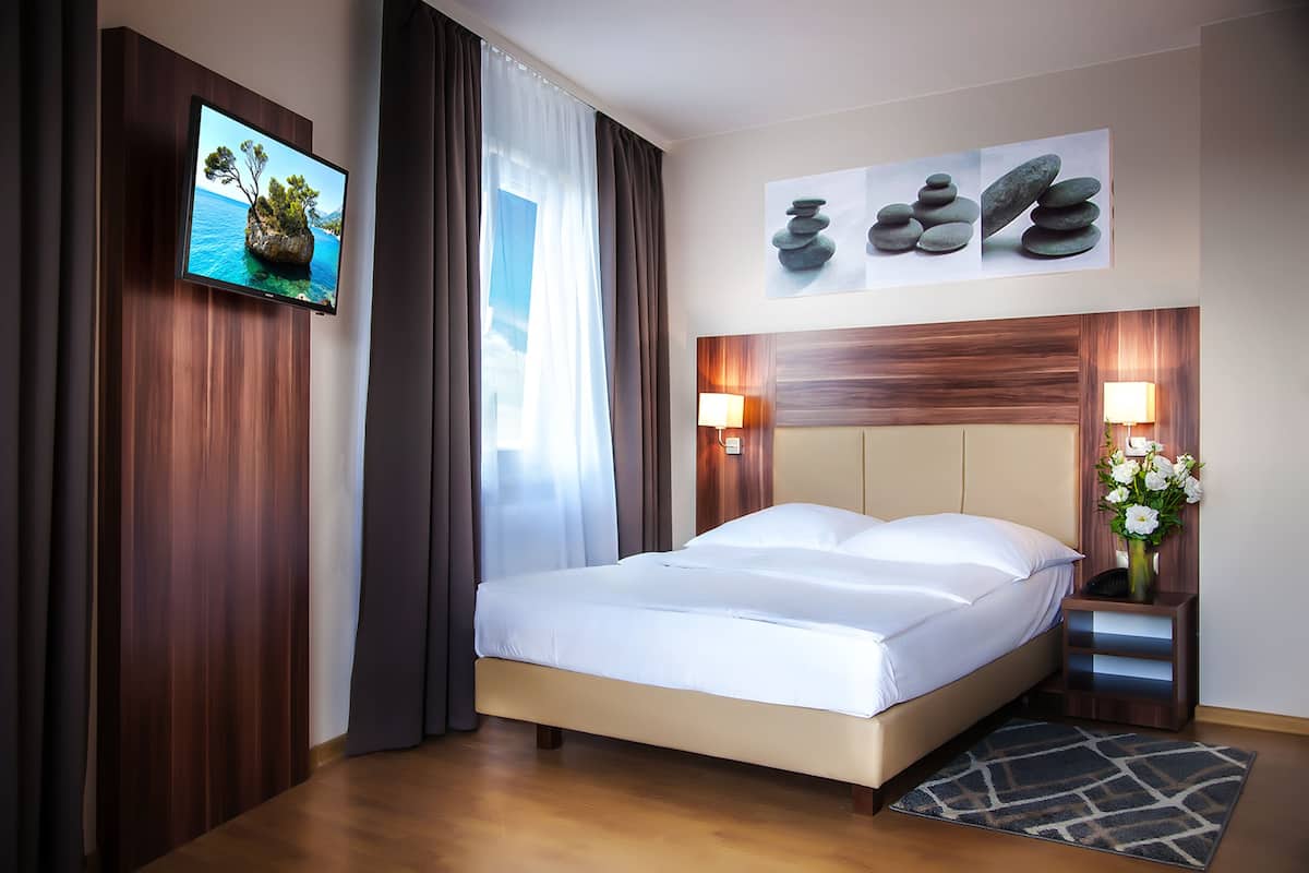 Quarto de hotel com cama, televisão fixada na parede, pintura de pedras e mesa de cabeceira com um vaso de flores.
