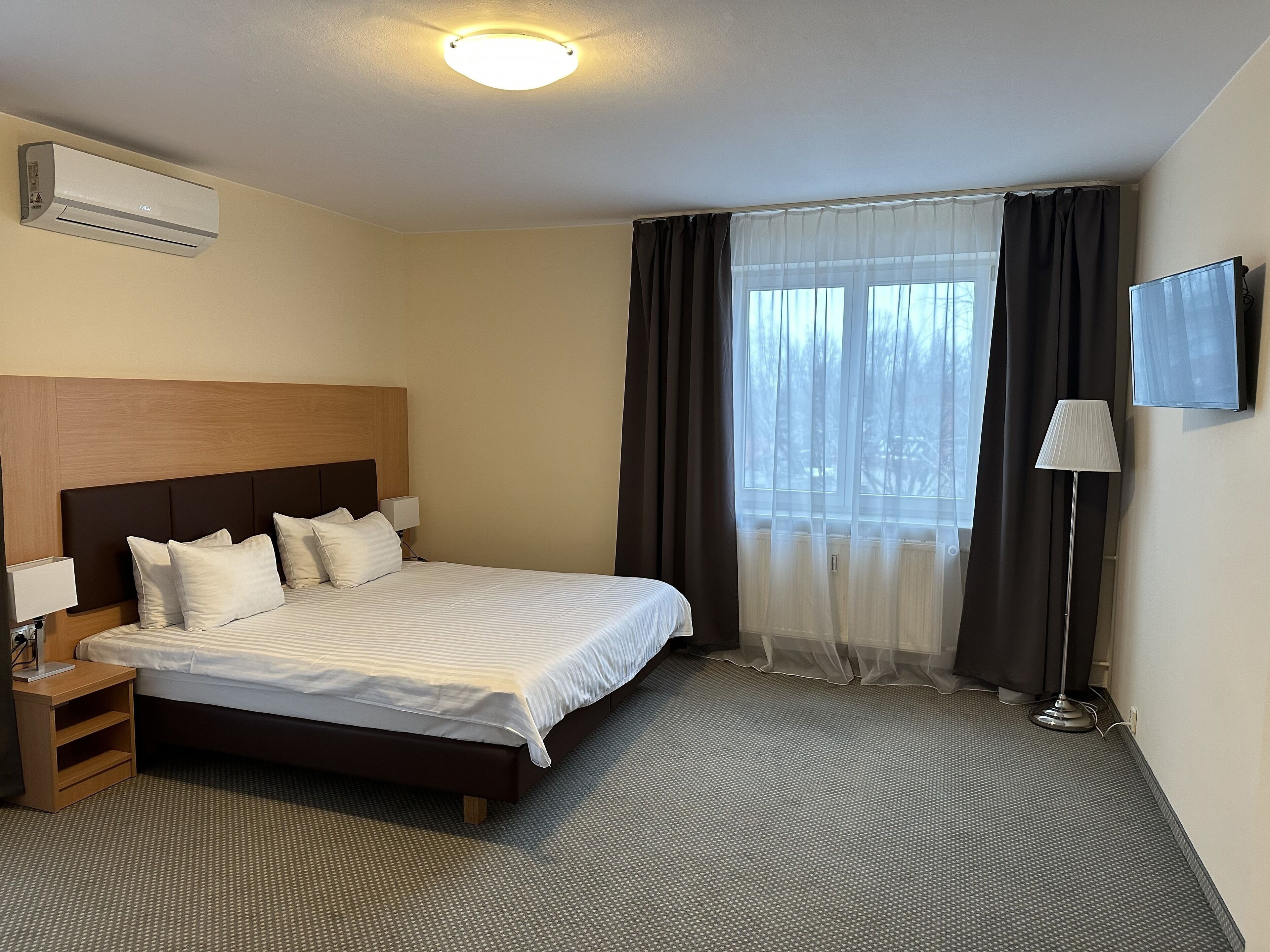 Ein Hotelzimmer mit Bett, Nachttisch, Lampe, Fernseher und einem Fenster mit Vorhängen.