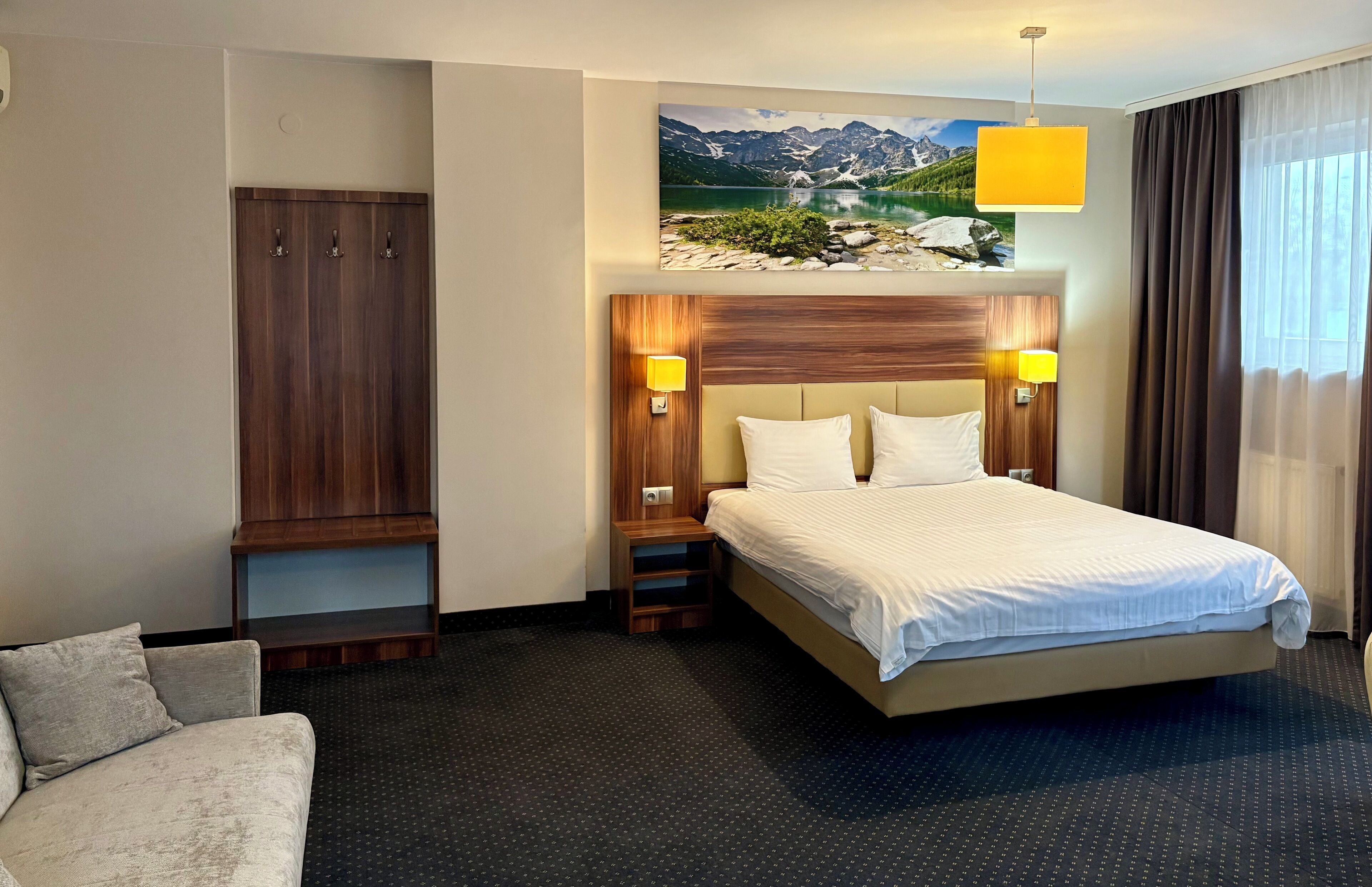 A hotel room with a large bed, a wardrobe, a sofa, a painting of a mountain landscape, and a window with curtains.