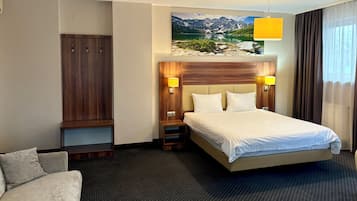 A hotel room with a large bed, a wardrobe, a sofa, a painting of a mountain landscape, and a window with curtains.