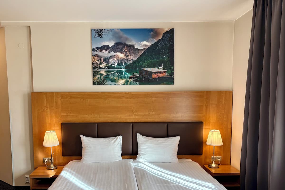 Quarto de hotel com uma cama grande, duas mesas de cabeceira, luminárias, uma cabeceira de madeira e uma pintura de paisagem montanhosa na parede.