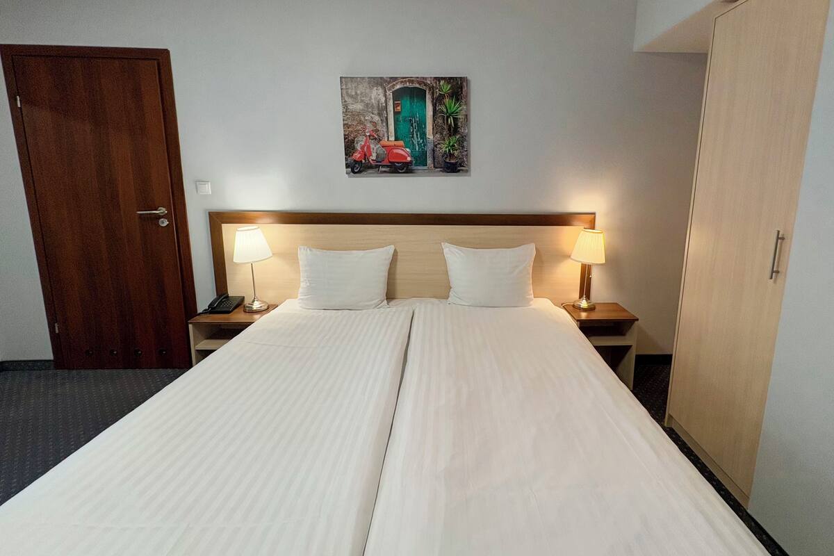 Quarto de hotel com uma cama grande, duas luminárias de cabeceira, uma cabeceira de madeira, uma porta, uma mesinha com um telefone e um quadro na parede.