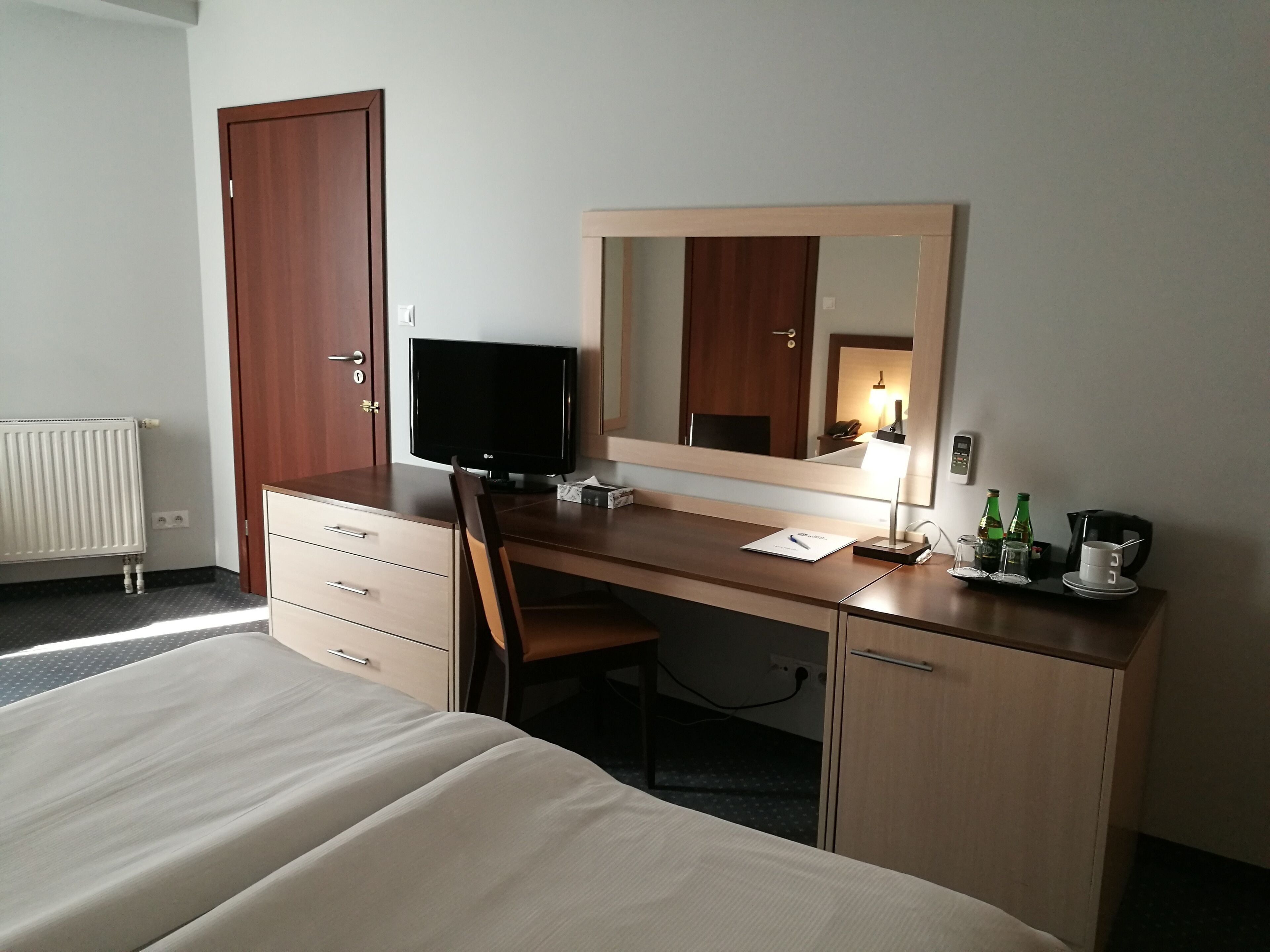 A hotel room with a bed, a desk with a computer, a mirror, a chair, and a bedside table with a lamp and some bottles.