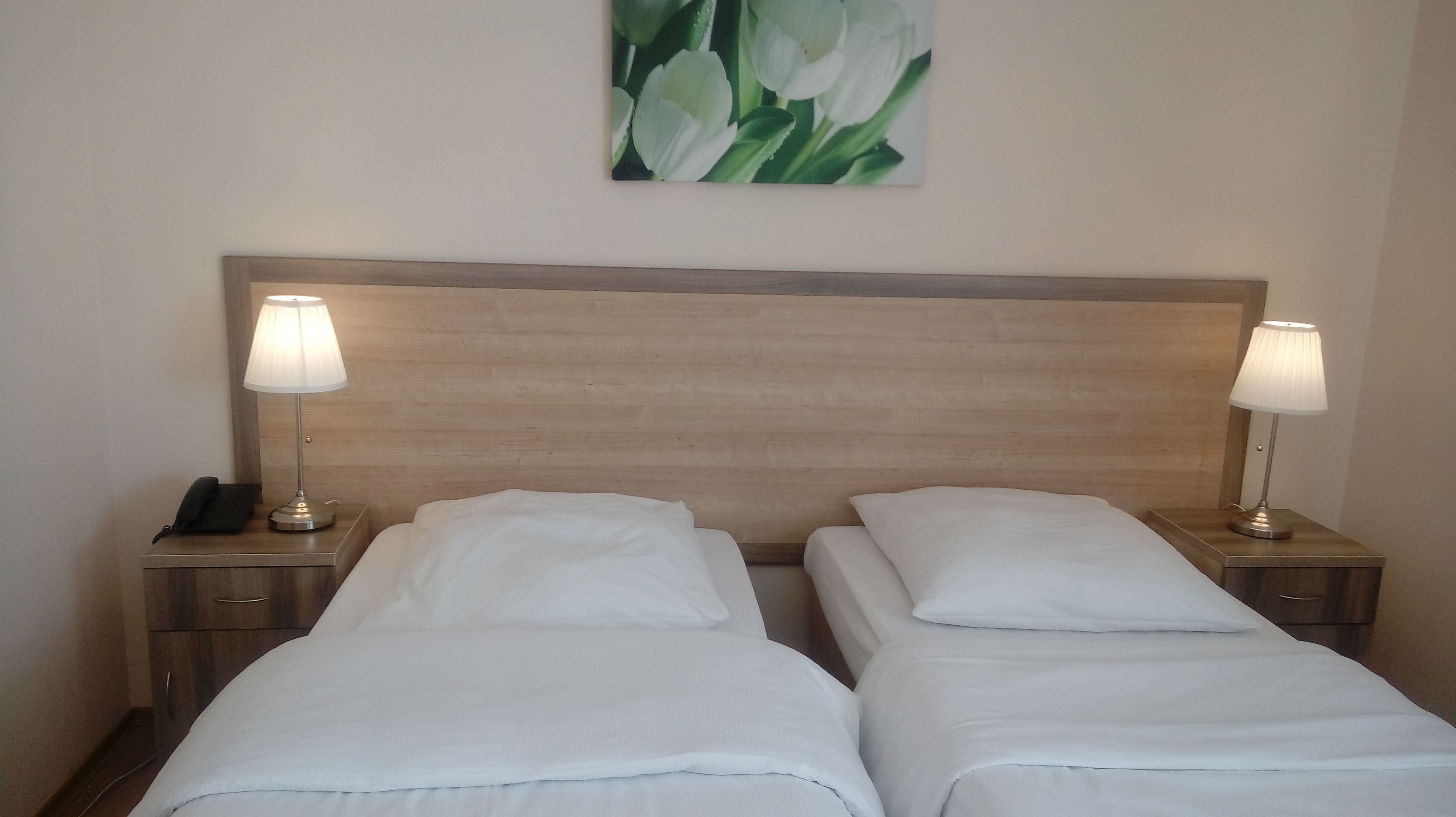 A double bed with white linens, two bedside lamps, a wooden headboard, a nightstand with a phone, and a painting of white flowers on the wall.