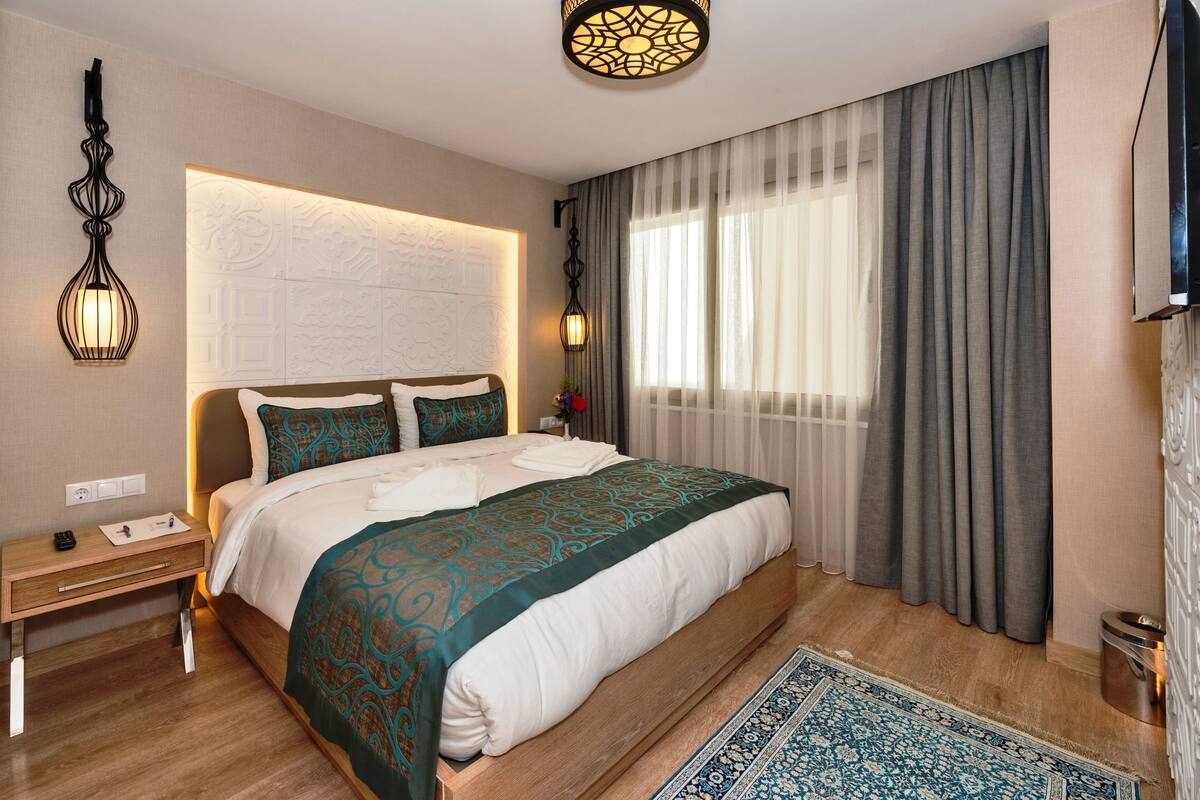 A hotel room with a large bed, a nightstand, a desk, a lamp, a window with curtains, and a patterned rug.