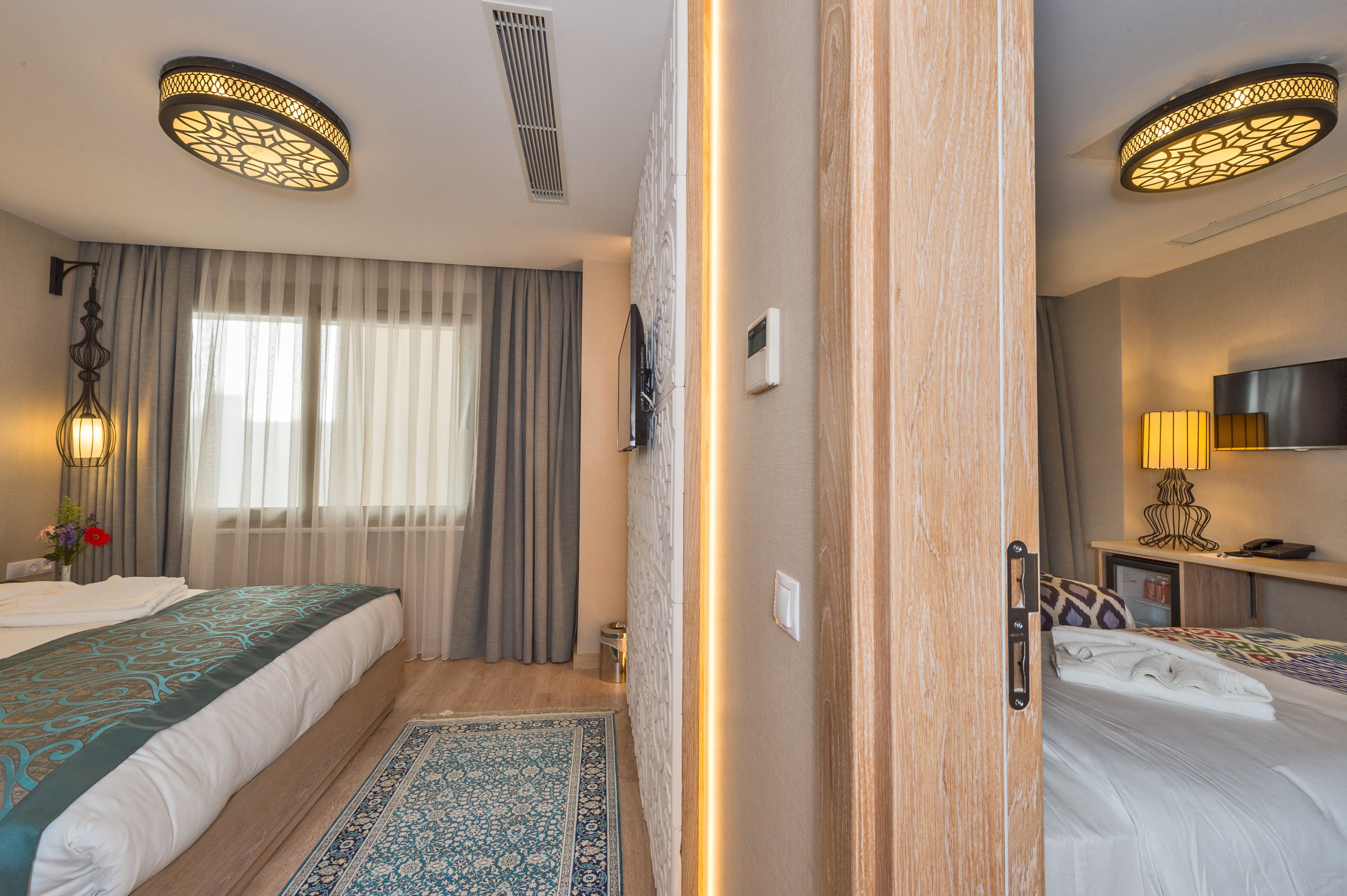 A hotel room with two beds, a large window with curtains, a wall-mounted TV, and a bedside table with a lamp.