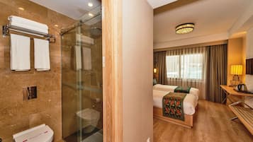 Une chambre d’hôtel avec une douche, des toilettes, un lit recouvert d’une couverture verte, une table de chevet avec une lampe et une fenêtre avec des rideaux.
