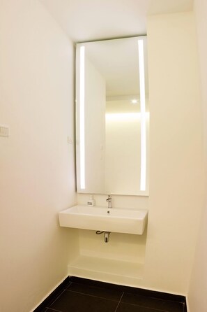 A small bathroom with a white sink, a mirror, and a black tiled floor. - Roomies Suites (George Town)