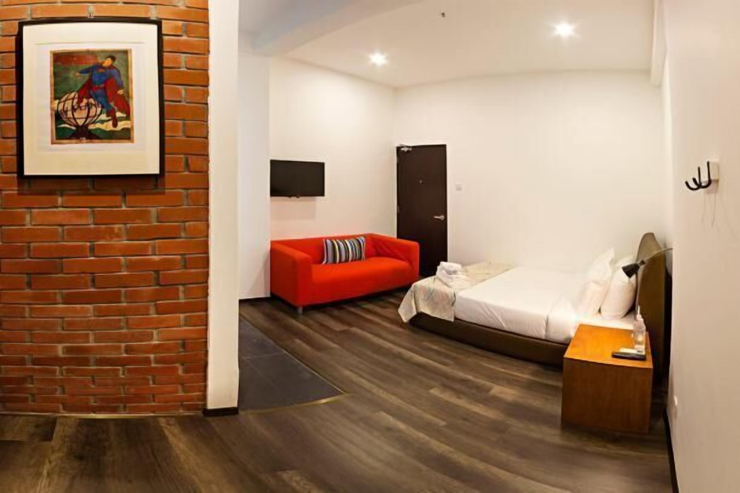 A hotel room with a brick accent wall, a red sofa, a bed with white bedding, and a flat-screen TV mounted on the wall.