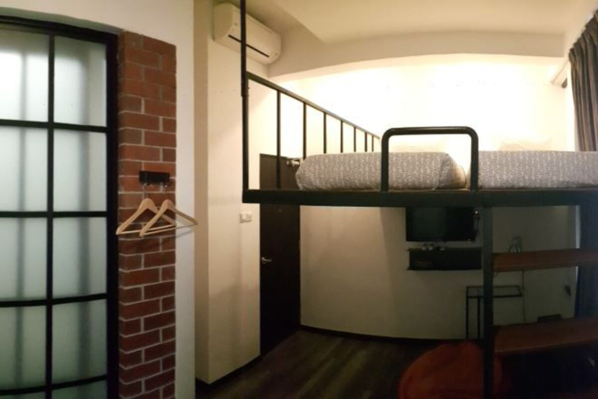 Une chambre avec des lits superposés, un mur en briques, une fenêtre avec des barreaux et un téléviseur placé sur le lit du bas.