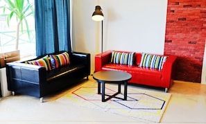 A living room with a black sofa, a red sofa, a small round table, and a floor lamp. - Roomies Suites (George Town)