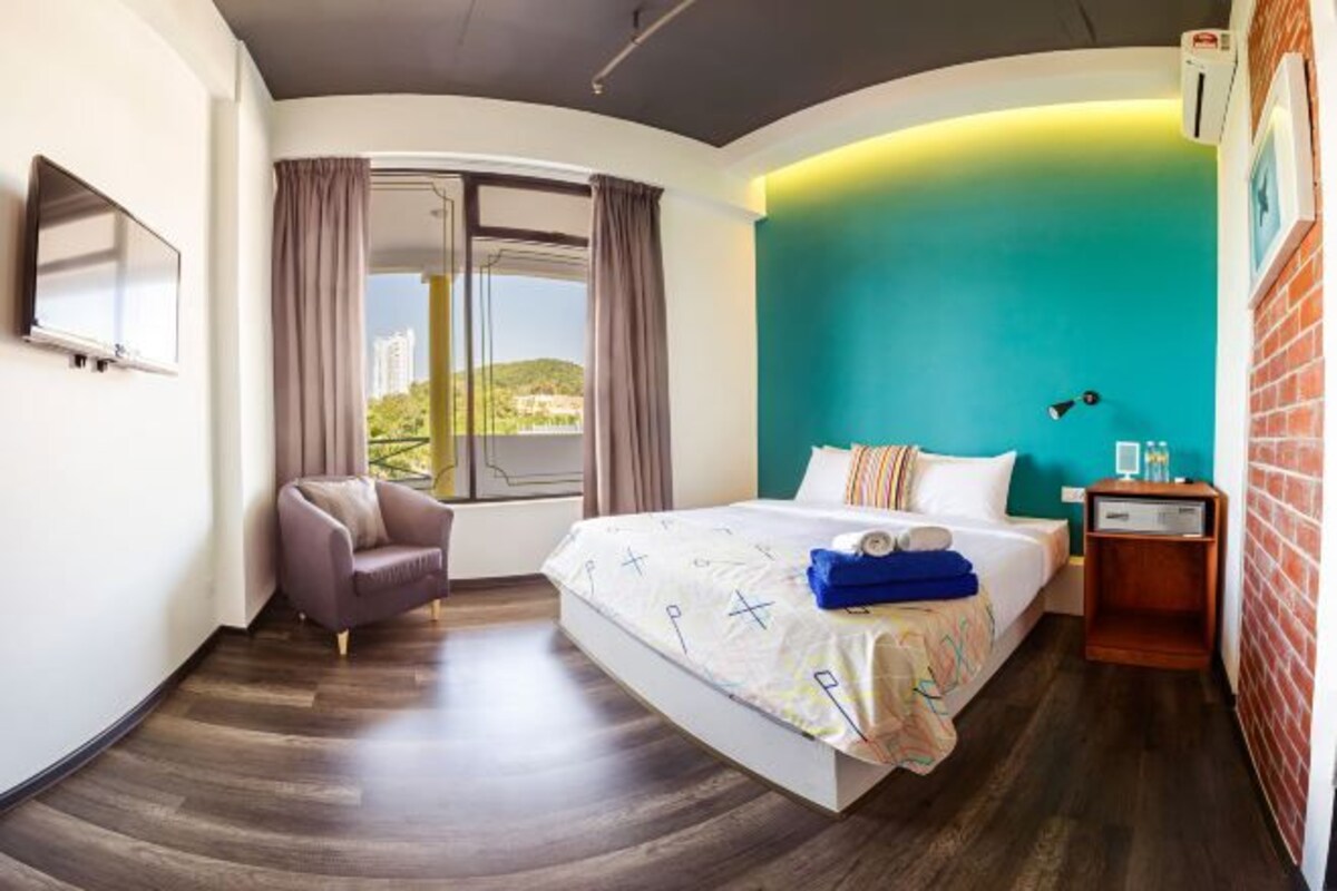 Une chambre d’hôtel moderne dotée d’un grand lit, d’un coin salon avec un fauteuil, d’un téléviseur fixé au mur et offrant une vue sur le paysage urbain par la fenêtre.