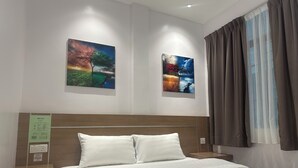 A hotel room with a bed, two paintings on the wall, a nightstand, and a window with curtains. - AST Hotel (Alor Setar)