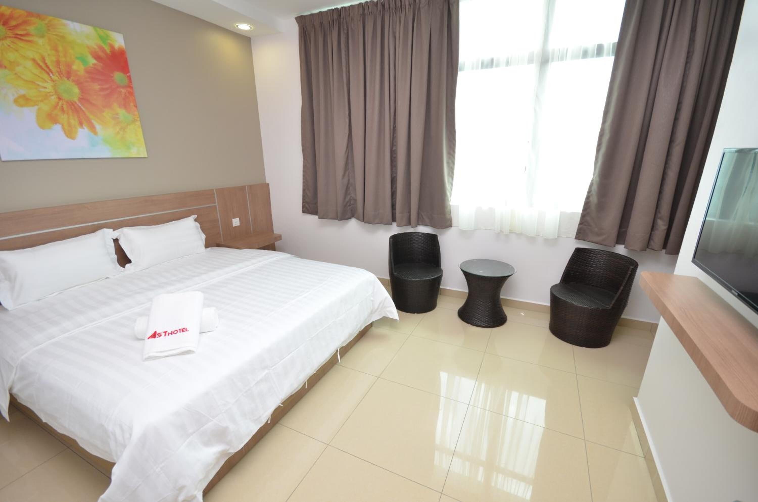 A hotel room with a bed, a TV, two wicker chairs, a small table, and a painting on the wall.