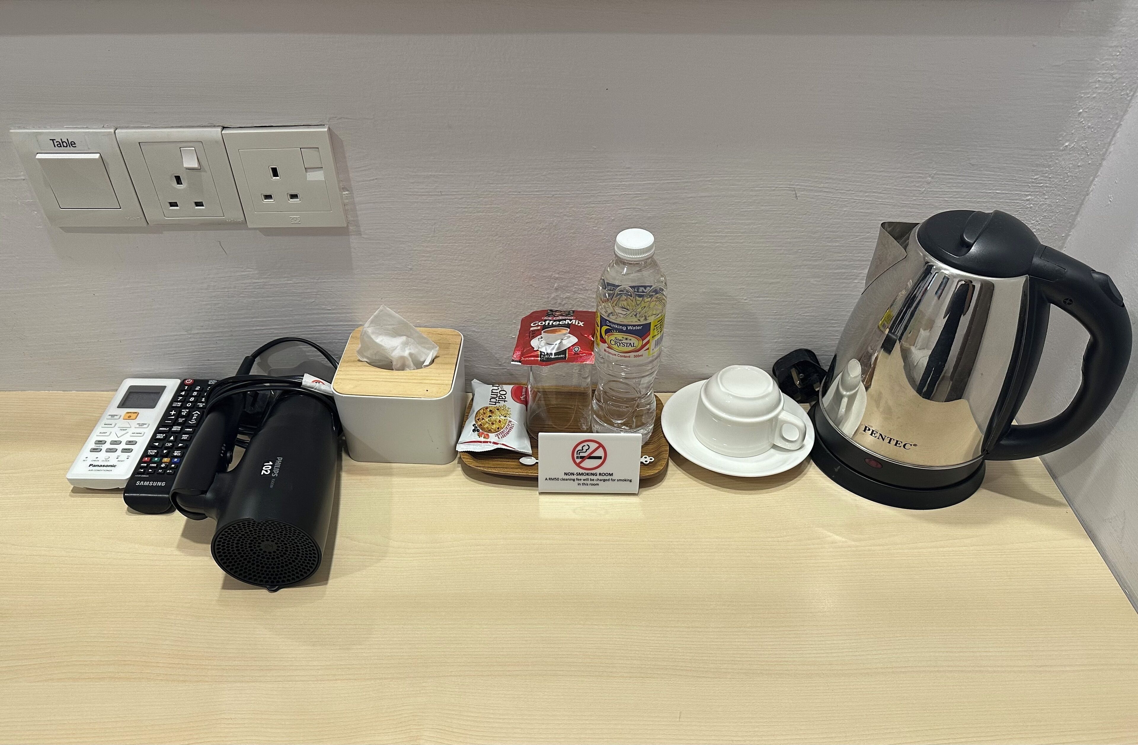 A hotel room with a table, a Panasonic phone, a Samsung charger, a black speaker, a tissue box, a packet of coffee, a bottle of water, a cup, a saucer, and a coffee maker.
