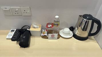 A hotel room with a table, a Panasonic phone, a Samsung charger, a black speaker, a tissue box, a packet of coffee, a bottle of water, a cup, a saucer, and a coffee maker.