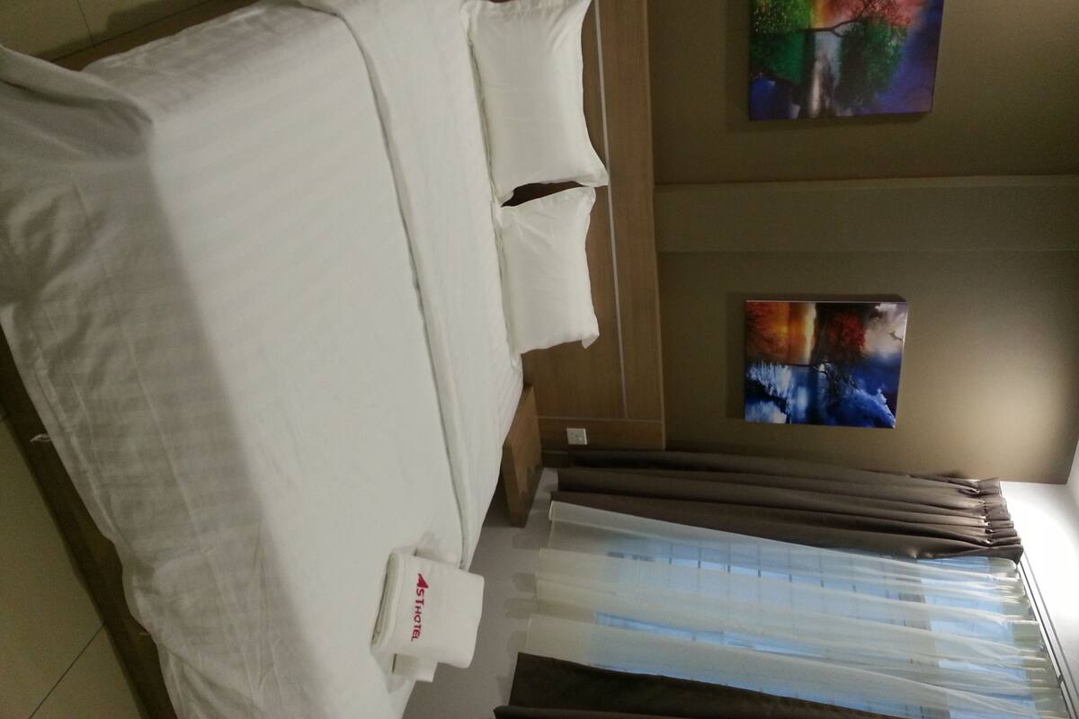 A hotel room with a bed, a window with blinds, and two paintings on the wall.