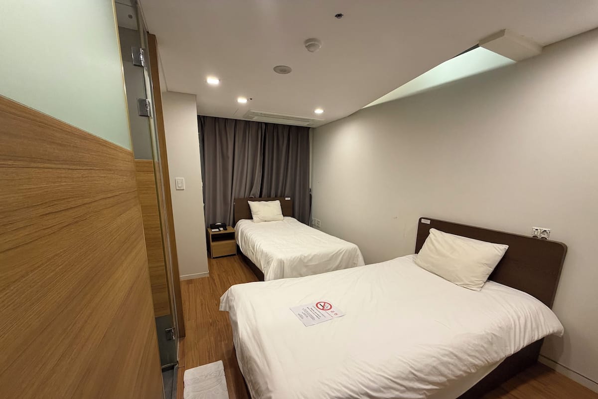 A hotel room with two single beds, a wooden floor, a small bedside table with a lamp, and a window with curtains.