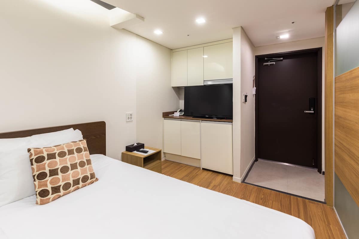 A hotel room with a bed, a bedside table, a TV, and a kitchenette.