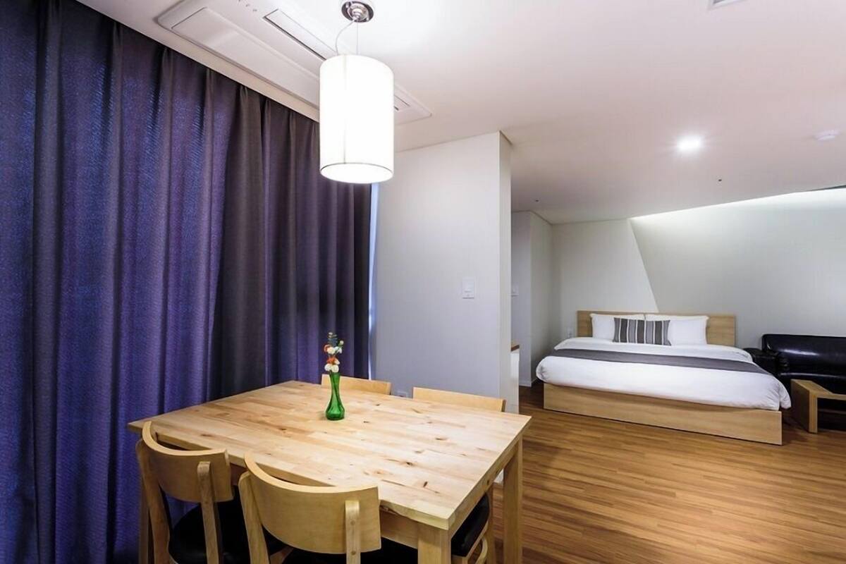 A hotel room with a wooden dining table, chairs, a bed with a white mattress and gray pillows, a black leather sofa, and a green vase with flowers.