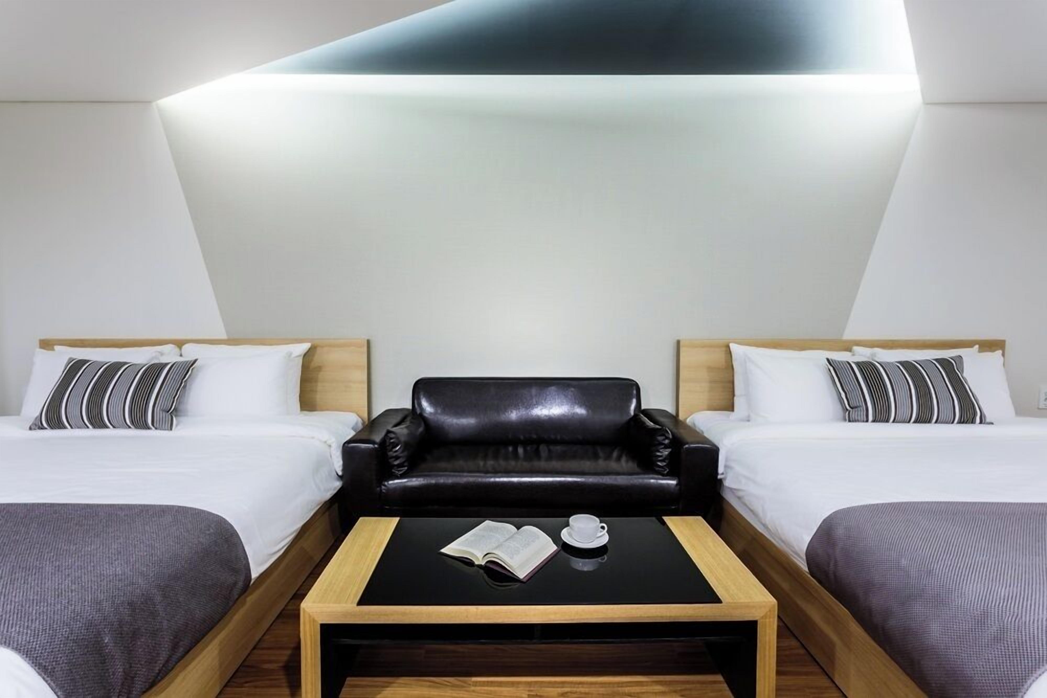 Et hotelværelse med to senge, en sort lædersofa, et lille træ sofabord med en bog og en kop, samt en moderne loftslampe.