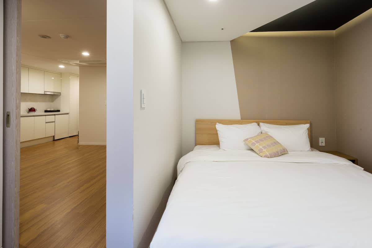 A hotel room with a wooden floor, a bed with white bedding, and a kitchen area in the background.