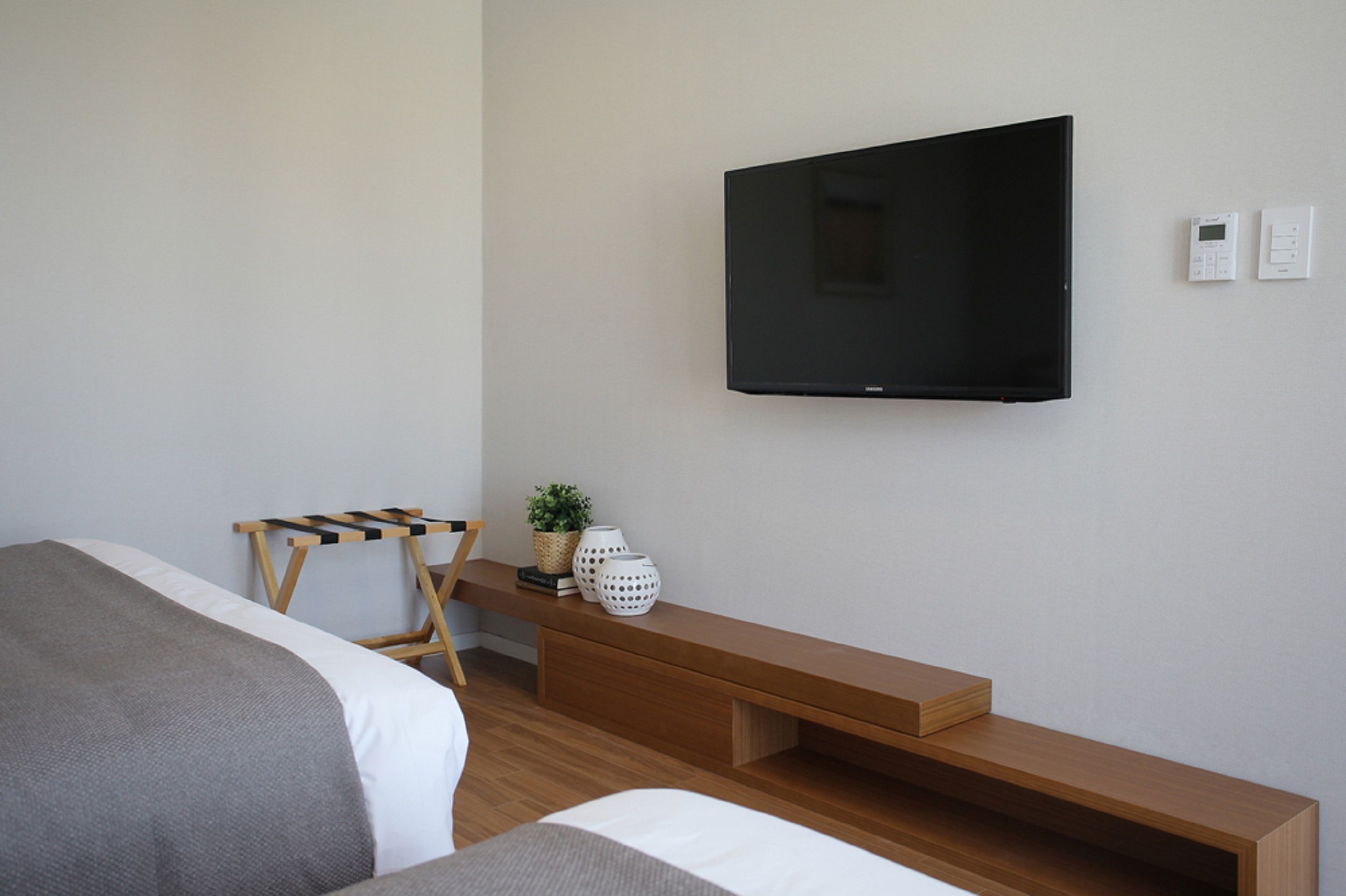 A modern hotel room with a large flat-screen TV mounted on the wall, a wooden bench, a small table with decorative items, and a bed with grey bedding.