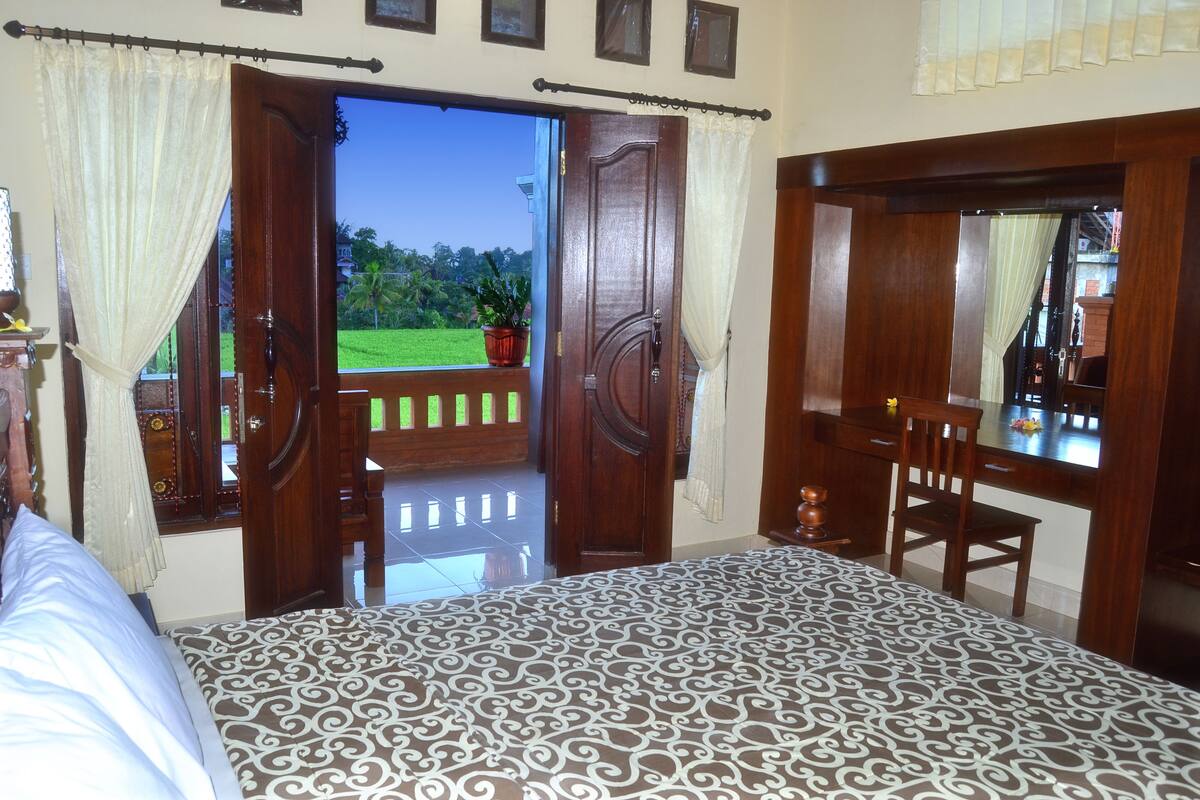 A bedroom with a patterned bedspread, a wooden desk, and a view of a green landscape through an open door.