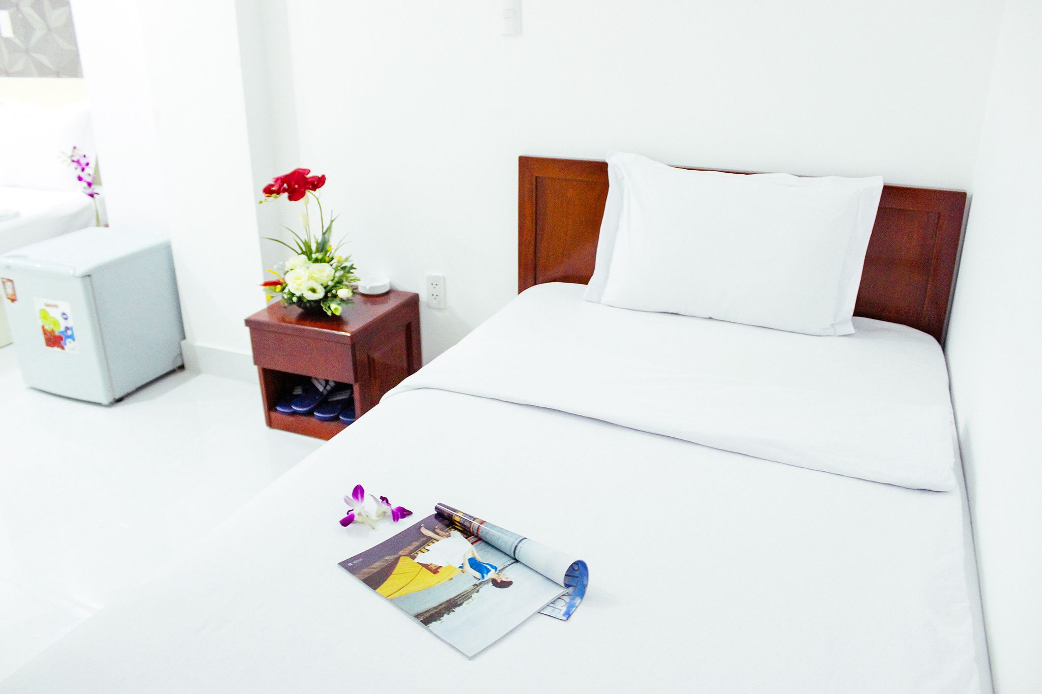 A hotel room with a neatly made bed, a bedside table with a vase of flowers, and a small refrigerator.