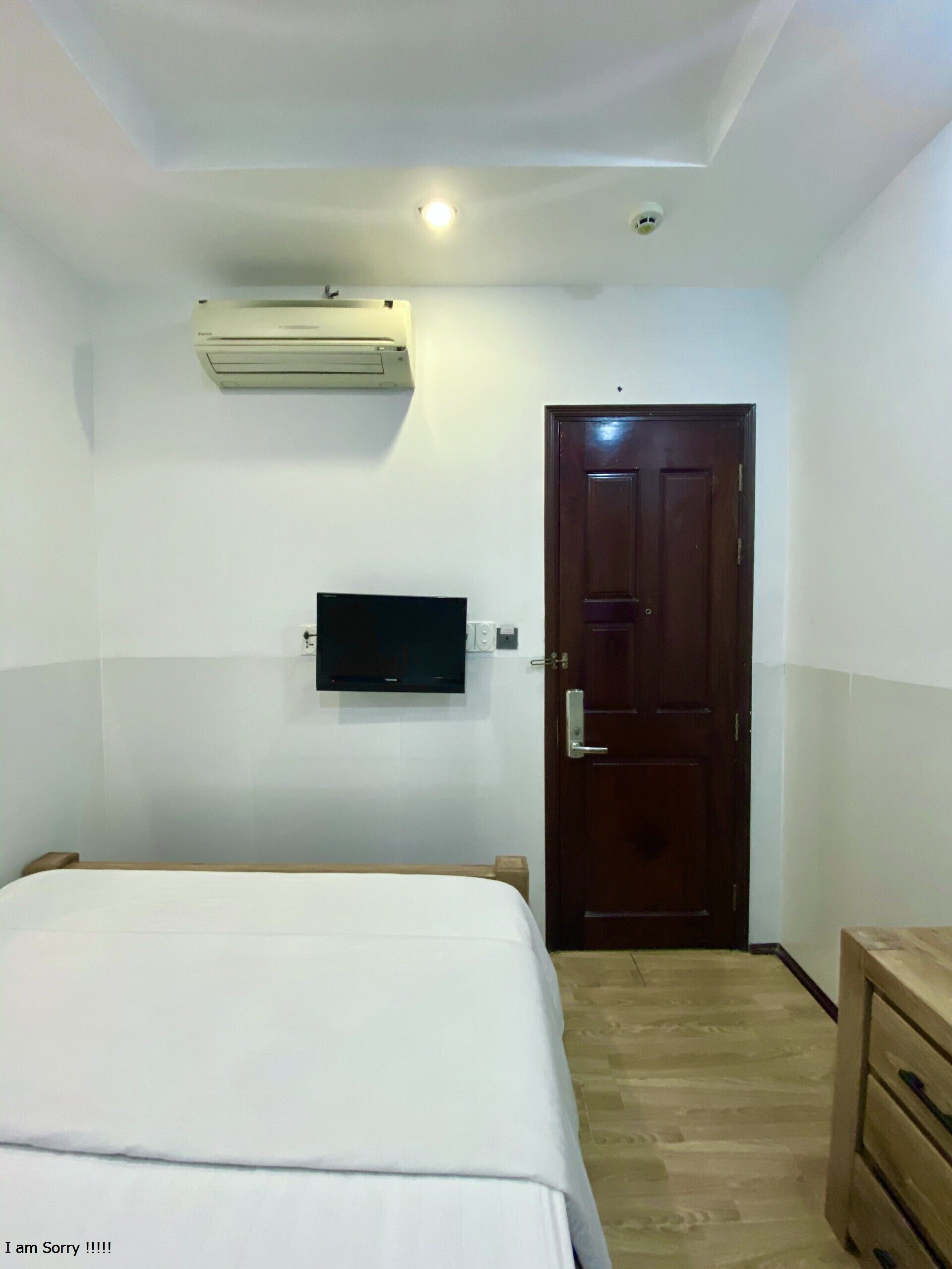 A small, neatly arranged bedroom with a bed, a wooden dresser, a television mounted on the wall, and a door.