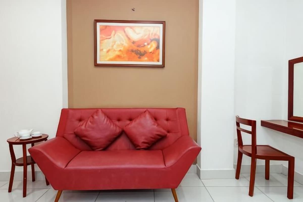 A red leather sofa with two cushions, a small round table with a white teapot and cups, a wooden chair, and a framed abstract painting on the wall.