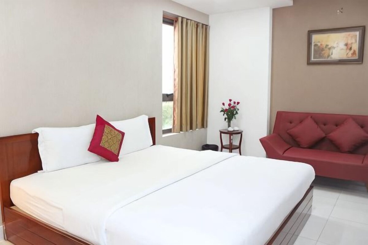 Habitació d'hotel amb un llit gran, un sofà vermell, una taula petita amb un ram i una finestra amb cortines.