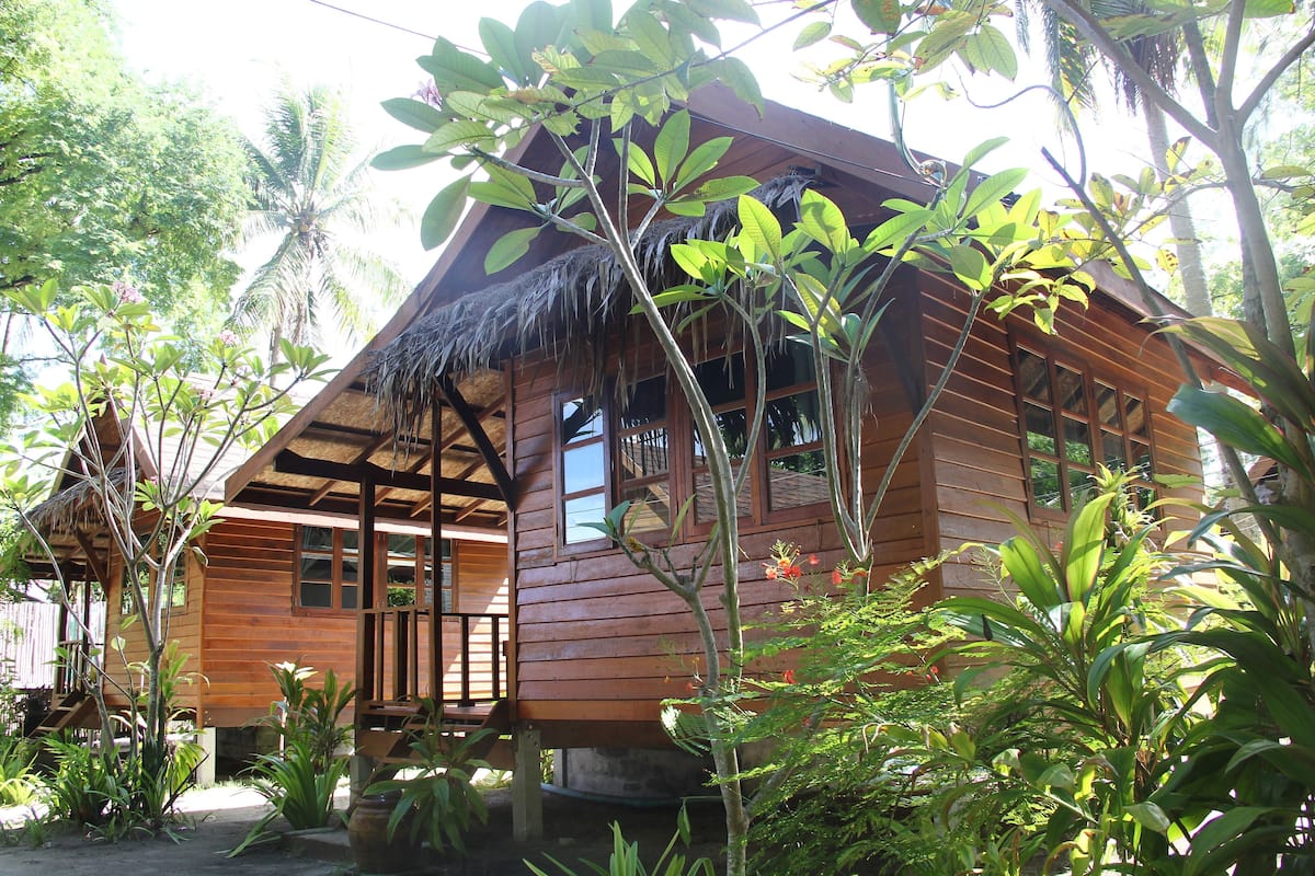 Uma casa de madeira com telhado de palha, rodeada por vegetação exuberante e palmeiras.