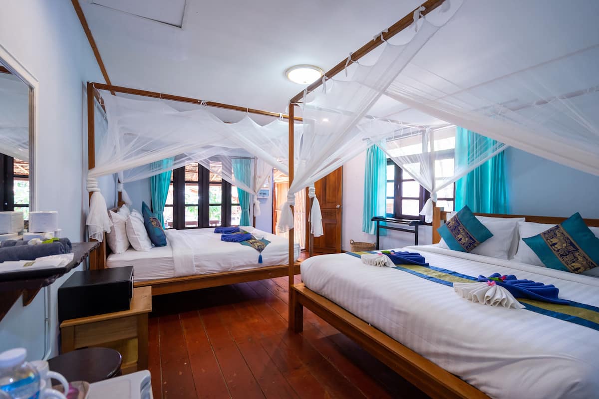 Quarto com duas camas, cada uma com dossel, cabeceira de madeira e roupa de cama branca. Há uma escrivaninha com um computador e uma cadeira, e uma janela com cortinas.