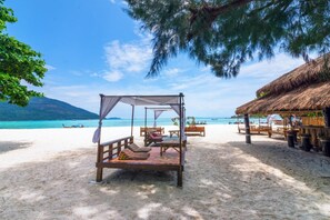 Une plage avec un abri à toit de chaume, un lit en bois doté d’un baldaquin, et une vue dégagée sur la mer et les montagnes en arrière-plan.