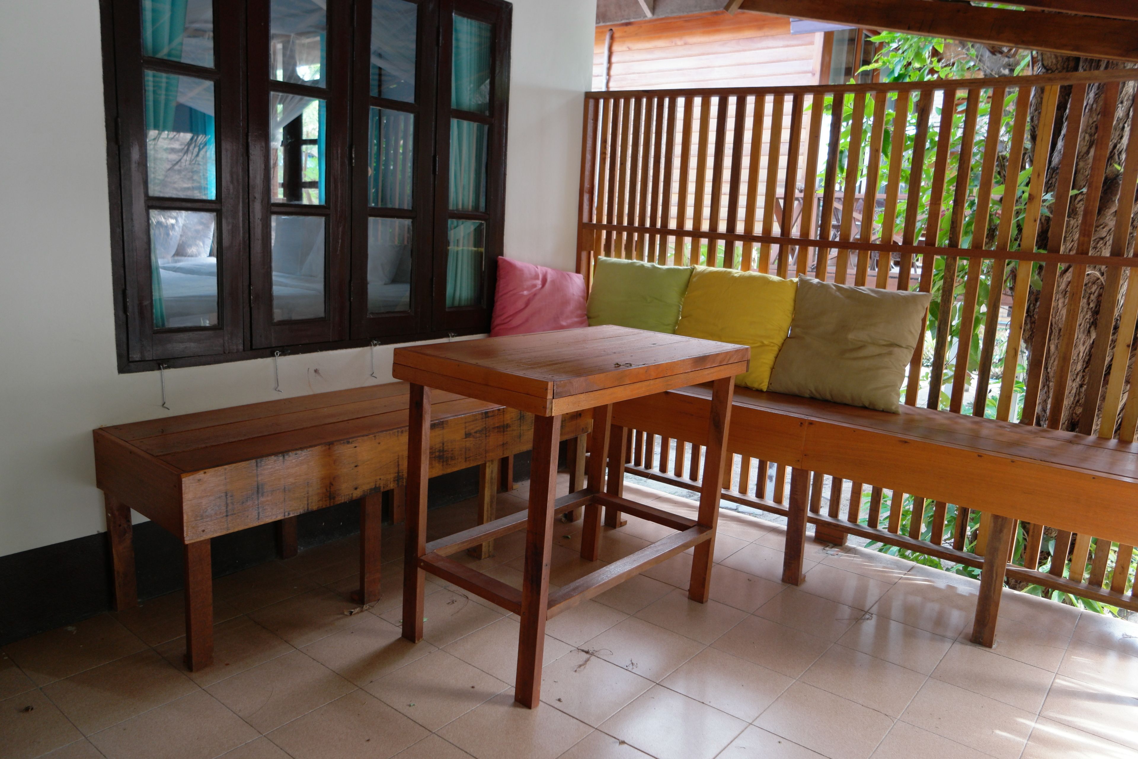A room with a wooden table and benches, a window with curtains, and a wooden partition with greenery outside.