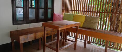 A room with a wooden table and benches, a window with curtains, and a wooden partition with greenery outside.
